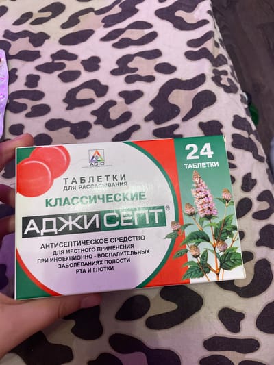 A person's hand is holding a box of medication. The box is predominantly white with green and red accents. The front of the box features the brand name "АДЖИ СЕПТ" in large, bold lettering, with "КЛАССИЧЕСКИЕ" above it. Below the brand name, it states "АНТИСЕПТИЧЕСКОЕ СРЕДСТВО ДЛЯ МЕСТНОГО ПРИМЕНЕНИЯ ПРИ ИНФЕКЦИОННО - ВОСПАЛИТЕЛЬНЫХ ЗАБОЛЕВАНИЯХ ПОЛОСТИ РТА И ГЛОТКИ". There is also an image of a flowering plant on the right side of the box, and a logo with "AGIO" and Russian text. On the upper right corner, it indicates "24 ТАБЛЕТКИ". The box is placed on a surface with a leopard print pattern. The image is a close-up shot, with the background blurred. The specific location within the city of Vaganovo, Russia is not discernible from the image.