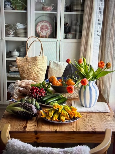This image, taken in Ros Na Rí, Ireland, captures a vibrant indoor scene, oriented vertically. A rustic wooden table or counter dominates the right side of the frame, featuring a strong wood grain pattern. Attached to the table is a light wooden hook holding a soft, fuzzy white object, possibly a garment or blanket.

The table is laden with an assortment of fresh produce and decorative items. A blue and white striped ceramic vase holds a bouquet of bright orange-red tulips, placed on a light-colored linen cloth. Beside the vase, a large oval dish, patterned in blue and white, overflows with delicate bright yellow zucchini blossoms. A bunch of dark green zucchinis, red radishes with their greens, and small gherkins are artfully arranged nearby. A distinct head of radicchio, with its deep purple and white leaves, rests at the forefront of the produce. Oranges or small pumpkins are partially visible behind the vase. A woven straw tote bag hangs over the edge of the table, and a transparent plastic bag containing unidentifiable items is nestled amongst the produce.

In the background, to the left, a white wooden cabinet or shelving unit with glass doors displays various dishware. Visible items include plates with a red/pink pastoral pattern, plain plates, teacups, saucers, and bowls, some matching the patterned plates, others in white or blue and white designs. Green decorative leaves are also placed within the shelves. Above the cabinet, a window with reflective glass shows indistinct objects or reflections, and horizontal blinds or shades are visible at the top.

The scene suggests a domestic setting, possibly a kitchen or dining area, showcasing fresh ingredients, implying a recent market visit, a harvest, or preparation for a meal. The arrangement creates a warm, inviting, and abundant atmosphere, characteristic of a cozy home environment. No people are visible, and no discernible text appears in the image. The time of day is not explicit, but natural light is present.