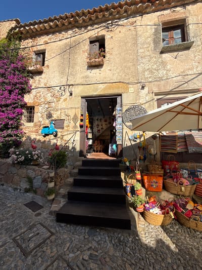 A sun-drenched scene unfolds in Tossa de Mar, Spain, showcasing the charm of a quaint Mediterranean village. The image focuses on the facade of a building with a rustic stone exterior, complemented by weathered walls and red-tiled roofing.
In the foreground, a narrow cobblestone pathway leads up a set of dark wooden steps, hinting at the steep terrain of the area. The pathway is adorned with various decorative objects, including woven baskets filled with colorful textiles, and an assortment of artisan crafts like decorative birds and a bright blue toy scooter. A vibrant display of fuchsia bougainvillea cascades down the stone wall on the left, adding a splash of color to the earthy tones of the building and pathway.
The building itself features several windows, some with wooden shutters and others with small planters overflowing with flowers. An open doorway reveals a glimpse into a shop, where more colorful items and textiles are displayed, suggesting a vibrant local market or boutique. A large, cream-colored umbrella provides shade over the entrance area, indicating fair weather suitable for outdoor browsing.
The sunlight casts sharp shadows across the scene, suggesting it is either morning or late afternoon. The overall impression is one of a picturesque and inviting village atmosphere, rich in local character and bathed in the warm glow of the Spanish sun. No people are visible, placing the emphasis on the setting and the objects for sale.