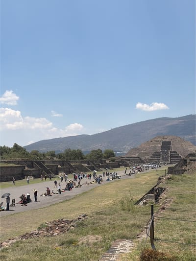 The image captures the Teotihuacan archaeological site in San Francisco Mazapa, Mexico. A large pyramid is visible on the right, and a mountain is visible on the left. A pathway runs between the pyramid and a grassy area, crowded with tourists. The sky is blue with a few white clouds. Trees are scattered along the pathway.