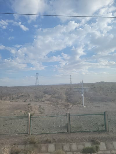 The image depicts a wide outdoor landscape with a focus on power lines and transmission towers against a backdrop of a vast, hazy sky filled with cumulus clouds. The scene is oriented vertically, suggesting it may have been taken from a high vantage point or with the camera tilted.

In the foreground, a section of a metal fence with green mesh and wooden framing runs across the ground, which is covered in gravel and sparse vegetation. Adjacent to the fence are paved areas or concrete structures.

Further into the scene, a large, lattice-style transmission tower stands prominently, supporting multiple power lines that stretch across the frame. In the distance, a more modern, single-pole utility structure with attached equipment is visible. The terrain is arid and mountainous, with the hills appearing barren and rocky, extending towards the horizon where they merge with the hazy sky.

The time of day appears to be daytime, given the bright illumination and the presence of distinct clouds. The weather seems to be fair but hazy, obscuring distant details. The location appears to be a somewhat remote, possibly industrial or infrastructure-heavy area within a desert or mountainous region. No people are visible in the image. The overall impression is one of infrastructure in a natural, rugged environment.