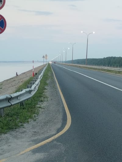 The image depicts a road on a clear day, likely in Zavolzhye, Russia, given the context. The road stretches into the distance, flanked by a guardrail and a body of water on the left. A single person is visible fishing by the water's edge. Streetlights line the right side of the road, and a dense line of trees forms a backdrop. Traffic signs are also present. The sky is a pale blue, with a few wispy clouds. The scene appears calm and uneventful.