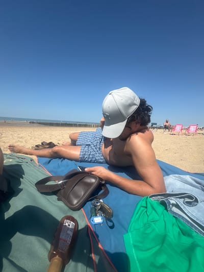 A person is lying on a blue beach towel on a sandy beach under a clear blue sky. The person is wearing blue patterned swim trunks and a light gray baseball cap that obscures their face. A brown leather fanny pack, keys, and a bottle of sunscreen are lying next to the person on the towel. In the background, there are other people and beach umbrellas. Wooden groynes can be seen along the shoreline. The location is Knokke-Heist, Belgium.