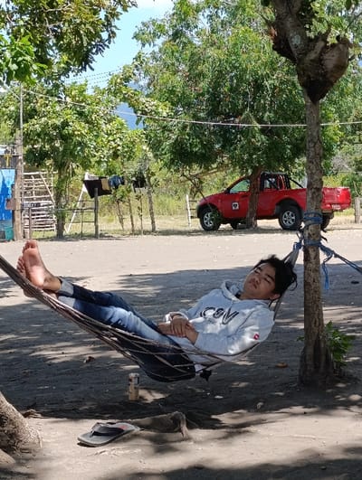 A young man is lying in a hammock, sleeping. He is wearing a white long-sleeved shirt with black lettering. His bare feet are visible, and he is wearing blue jeans. He is laying in a hammock under a large tree, which is on a sandy beach. The hammock is hanging between the tree and another support beam. A red truck is parked in the background. The scene is in Kecamatan Banyuputih, Indonesia. The coordinates are latitude: -7.757, longitude: 114.375.