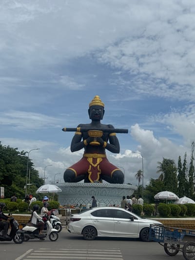 The image shows a large, dark statue of a seated figure wearing a gold headdress and elaborate gold and red clothing. The figure holds a golden bowl in its hands and has a long, dark cylindrical object resting on its shoulders and held by its arms. The statue is positioned on a large, ornate blue and white base. In the foreground, there is a street with a white sedan driving on it. Several people are visible around the statue, some standing near it and others on motorcycles. There are also white umbrellas with a "BM" logo scattered around the base of the statue. The environment appears to be a public square or roundabout in Battambang, Cambodia, with trees and streetlights visible in the background. The sky is cloudy.