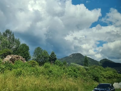 The image shows a landscape with rolling green hills and mountains under a dramatic sky with both dark storm clouds and bright white cumulus clouds. In the foreground, there is lush green vegetation, including tall grass and bushes. To the left, a large pile of cut and stacked lumber is visible. A dark-colored SUV is parked on the right side of the frame, partially obscured by the vegetation. The scene is likely in or near St. Peter ob Judenburg, Austria, given the mountainous terrain and prevalent greenery often found in Austrian rural areas. There are no people or specific actions depicted in the image, making it a static, environmental scene.
