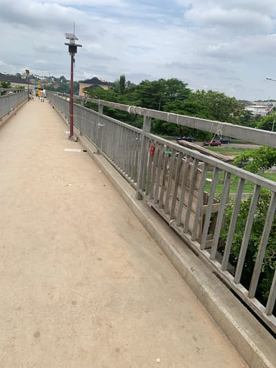 The image depicts a pedestrian overpass in Jiru, Nigeria. A concrete walkway runs diagonally from the bottom left to the top left of the frame, bordered by a metal railing on the right. Several people, seemingly pedestrians, are visible on the walkway, walking away from the camera. One individual is seated on the ground near the railing. A lamppost with a solar panel is mounted on the walkway, and another similar structure is visible in the distance. Beyond the railing, a busy road with several cars is visible, as are numerous trees and buildings. The sky is overcast with clouds.