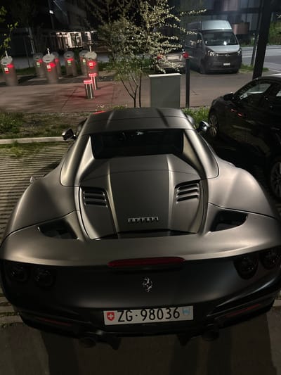 The image displays a matte dark gray Ferrari sports car parked outdoors at night, viewed from the rear-right. The car's sleek lines, pronounced rear fenders, and multiple air vents on the engine cover are prominent. The word "Ferrari" is visible in chrome lettering on the trunk lid, accompanied by the iconic prancing horse emblem. The rear of the vehicle features quad exhaust tips and a red horizontal taillight bar.
A Swiss license plate, "ZG - 98036," is clearly displayed on the back of the Ferrari, indicating registration in the Canton of Zug. The Swiss flag and the Zug canton emblem are also visible on the plate.
To the left of the Ferrari, a series of modern, cylindrical bollards with illuminated red tops are arranged, suggesting an access control point. Beyond these bollards, trees or bushes with light-colored blossoms add a touch of nature to the scene. In the background, other vehicles are parked, including a dark SUV or sedan partially obscured by the Ferrari, a light-colored commercial van, and another dark sedan. The setting is a paved outdoor area, possibly a private driveway or parking lot, dimly lit by artificial lights, indicating nighttime or late evening. The location is contextualized as Dübendorf, Switzerland.