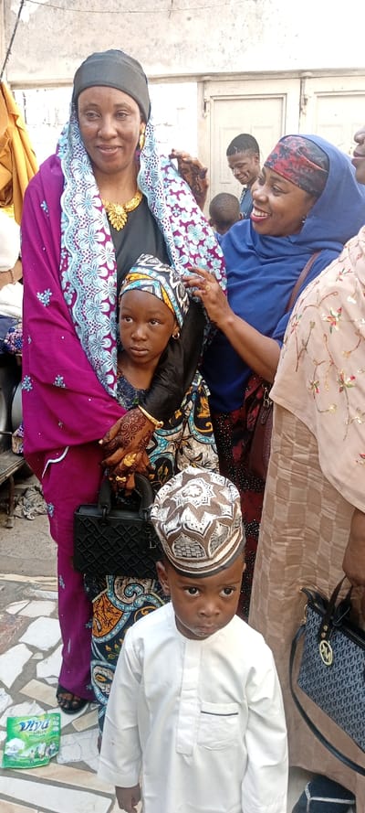 The image depicts an outdoor social gathering in Dan Maliki, Nigeria, centered around five individuals in the foreground and midground, with additional figures partially visible in the background.
In the foreground, a young boy with dark skin stands facing forward, looking directly at the viewer with a serious expression. He wears a white, long-sleeved tunic with a small left chest pocket outlined in black, and a light-colored, intricately patterned kofia (cap) with brown and white designs. To his left, a green plastic pouch labeled "Viva Plus" lies on the patterned tiled ground.
Behind the boy and to the left, a woman with dark skin stands, holding a young girl. The woman wears a dark head wrap, large gold hoop earrings, a prominent gold necklace, and a magenta/purple garment with a blue and white patterned shawl. Her hands are adorned with intricate henna patterns and gold rings and bangles. She holds a small, black quilted handbag with a top handle. Her facial expression is a subtle smile.
The young girl held by this woman has dark skin and wears a patterned head wrap and a dark top. She looks slightly to her left with a neutral, perhaps pensive, expression. Small gold hoop earrings are visible in her ears.
To the right of the central woman and child, another woman with dark skin smiles broadly, looking towards the girl. She wears a blue headscarf and a blue garment, and her right hand, adorned with henna, is positioned near the girl's head wrap, possibly adjusting it.
Partially visible on the far right is a fourth woman, wearing a light beige or tan garment with floral embroidery and holding a black Michael Kors handbag with a patterned strap. In the background, behind the smiling blue-clad woman, a man with a short haircut smiles, and another child is partially visible. The background features a light-colored, textured wall and a closed double-leaf white door. The ground is paved with irregular light-colored tiles. The scene is illuminated by natural light.