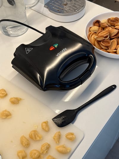 The image displays a kitchen countertop scene in Башкултаево, Russia, focused on the preparation of Oreshki cookies. A black Viconte electric appliance, designed for baking, is prominently featured, displaying both red and green illuminated indicator lights and a warning symbol for heat. To the left of the appliance, a clear glass pitcher filled with water is visible. To the right, a white bowl holds a quantity of baked, golden-brown, shell-shaped cookies. In the foreground, a white cutting board is populated with multiple small, unbaked portions of pale yellow dough. A black spatula with a perforated handle lies on the countertop near the cutting board. A segment of a larger white kitchen appliance with a metallic grater is partially visible in the upper background. No individuals are present in the frame.