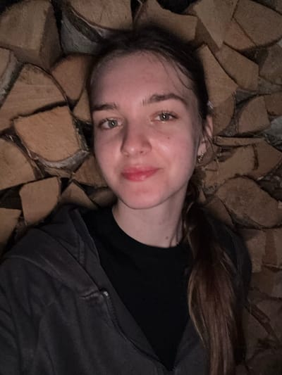 The image is a close-up, overhead shot of a young woman with long, brown hair tied back in a ponytail. She is wearing a dark gray hooded jacket over a black t-shirt. The woman is positioned in front of a backdrop of stacked firewood. The lighting is dim, casting shadows and highlighting her features. There are no discernible elements of a city, or specifically Nizhnie Mully, Russia, visible in the image. The focus is on the woman and the firewood.