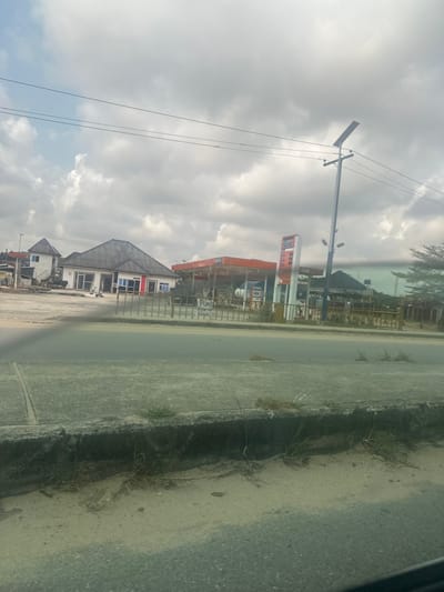 This image depicts a roadside scene in Elimgbu, Nigeria. The foreground shows an asphalt surface leading to a concrete curb, beyond which is a strip of sandy ground with sparse dry grass and weeds. Across an asphalt road, a low metal fence spans the middle ground. Behind the fence, to the left, is a single-story white building with a dark, sloped roof and multiple windows, some featuring red accents. To its left, a multi-story building with a pointed roof is partially visible. To the right of the white building, an APL-branded gas station is present, characterized by an orange canopy covering fuel pumps and a tall white and blue vertical sign displaying prices. A utility pole fitted with a solar panel stands near the gas station, supporting multiple overhead power lines that extend across the upper frame. A handwritten sign on the metal fence reads "PUMP" followed by indistinct text. The sky is largely overcast with scattered clouds. No discernible individuals or active vehicles are present in the immediate depicted area.