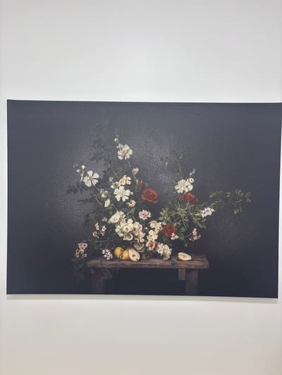 This is a horizontal, close-up, medium shot of a framed canvas art piece hanging on a white wall. The canvas depicts a still life of a floral arrangement with fruit on a wooden table, set against a dark, textured background. The bouquet is rich with various flowers, including red roses, white roses, and smaller pink and white blossoms, interspersed with green foliage. Two whole pears and one halved pear are arranged on the rustic wooden table in the lower portion of the canvas. The lighting is dim, creating a dramatic and moody atmosphere typical of classical still life paintings. The canvas appears to be unframed and stretched taut. The white wall shows subtle texture. The location of this artwork is in the city of Berchem-Sainte-Agathe - Sint-Agatha-Berchem, Belgium.