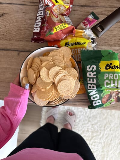 In this top-down shot, a spread of snacks is laid out on a wooden table and a fluffy white rug, suggesting a casual indoor setting. The primary objects are various snack bags and a bowl of round, crispy chips. Among the visible snack packaging are "Bombar" protein chips in green and yellow bags, one featuring mushrooms and the other with bold "Bombar" branding. Another bag, red and black, advertises "Bacon & Paprika" flavor. A smaller pink package with "Raspberry Raf" is also present. The chips themselves are piled high in a white bowl with a black rim.
A person's hands, one wearing a pink long-sleeved shirt and the other holding the bowl of chips, are visible in the foreground, indicating someone is about to partake in the snacks. The person's fingernails are painted light blue with white polka dots. In the background, on the white rug, another person's legs are visible, wearing black pants and light-colored slippers, suggesting they are also present. The scene appears to be set indoors during daylight, with the overall mood being relaxed and informal. The text on the packaging provides details about the snack brands and flavors.