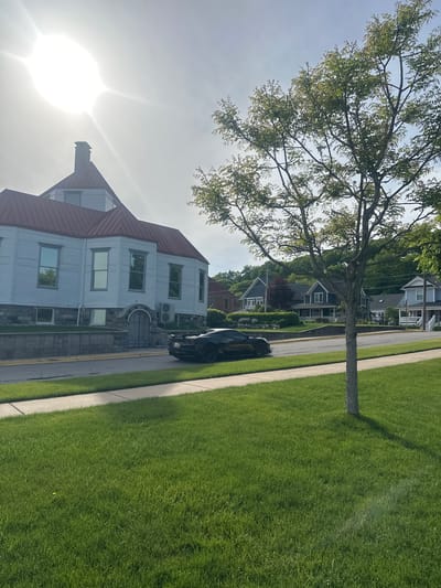 A street scene in Harbor Springs, Michigan, USA, on a sunny day. The image features a light blue building with a red roof and chimney on the left, appearing to be an institutional or historical structure. In front of it, a black sports car is parked on the street, with its rear visible. The street is lined with a sidewalk and a well-maintained green lawn. In the background, across the street, are several residential houses with pitched roofs, varying in color from dark blue to light blue, set amidst trees and landscaping. A mature tree with light-colored bark and green leaves stands in the foreground on the right, casting a shadow on the lawn. The sun is bright and high in the sky, causing some lens flare.