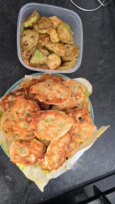 Two dishes of food are presented on a dark gray countertop. In the upper portion of the image, a clear plastic container holds a pile of sliced, pan-fried zucchini. The zucchini slices are round and vary in color from light brown to green, with some exhibiting darker, crisped edges. Below the container, a light blue plate is filled with a stack of golden-brown fritters. These fritters appear to be made from a batter with visible flecks of green herbs or scallions. The plate is lined with a white paper napkin with yellow trim. The scene is indoors and appears to be in a kitchen environment, though specific details of the location within Butor, Moldova are not discernible from the image alone.