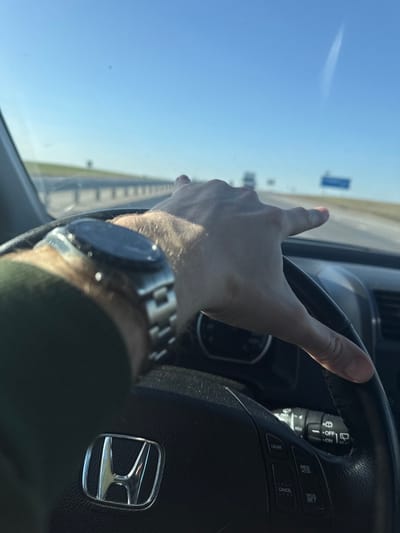 The image displays a first-person perspective from the driver's seat of a Honda vehicle in Абухаўскі сельскі Савет, Belarus, while in motion. The driver's right hand, wearing a metallic smartwatch on the left wrist, grips the upper right portion of the black leather steering wheel, which features a silver Honda emblem in the center and cruise control buttons on the right spoke. The driver is clad in a dark green long-sleeved garment. Ahead, a multi-lane highway stretches into the distance under a clear blue sky, containing a faint aircraft contrail. A metallic guardrail lines the left side of the road, and distant vehicles along with a blue rectangular road sign are visible further down the highway. The scene suggests a sunny day with unimpeded forward visibility.