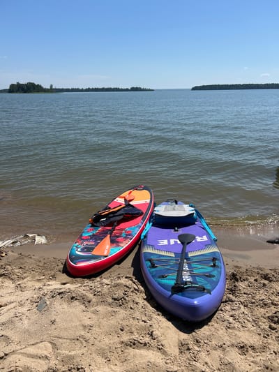 Two stand-up paddleboards and paddles are situated on a sandy beach next to a body of water under a clear blue sky. The paddleboard on the left is red and orange with a colorful graphic design, featuring a life jacket on top. The paddleboard on the right is purple with a blue and yellow graphic design and a black paddle. In the background, a tree-lined horizon separates the water from the sky. The scene appears to be a calm day at a lake or reservoir, possibly in the city of Borovoye, Russia, which is known for its natural beauty and bodies of water.