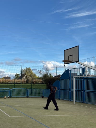 An outdoor multi-sport court, identified as being in Niederhausbergen, France, is depicted under a bright blue sky with scattered white clouds and multiple horizontal contrails. The court surface is light green asphalt with white and blue demarcation lines. A single individual, appearing male, is standing on the court with his back partially to the camera, hands on his hips, and facing left. He is dressed in black attire: a t-shirt, pants, and shoes. To the left of the individual, two deflated soccer balls rest on the court. To the right, a basketball hoop with a white rectangular backboard and an orange rim is visible, supported by a white pole. A blue, slatted barrier structure, which includes a white metal goal frame, is located behind the individual and the basketball hoop. The court is enclosed by a tall green wire mesh fence, with lower blue metal barriers lining its base. Beyond the fence, a grassy field contains several trees, including one with yellowish-green foliage, and a brown animal, possibly a deer, grazing. In the far background, residential buildings with sloped roofs are partially visible, along with a field of tall, dried stalks, likely corn.