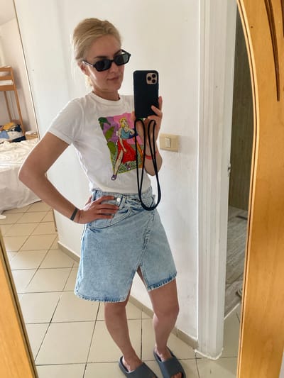 A fair-skinned female is posing in front of a mirror, holding a black smartphone with a black strap attached. She is wearing dark sunglasses, a white t-shirt with a graphic design, and light blue denim shorts. She is also wearing gray sandals. The environment appears to be the interior of a room, possibly a bathroom, with light-colored tiled floors and white walls. A light switch is visible on the wall. In the background, there is a partially visible bed and a wooden rack with clothing. The reflection in the mirror reveals additional details of the room. Based on the given location, this scene is taking place in Monastir, Tunisia.