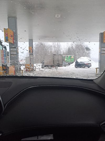 A snowy scene is viewed from inside a vehicle. Several trucks are lined up in a parking lot, visible through the windshield, which is covered in droplets. A grey building stands to the left of the trucks. The interior of the car is visible on the right, including a dark dashboard.