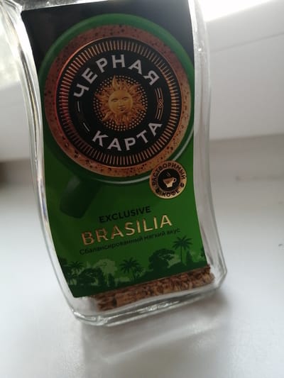 A glass jar containing coffee granules is positioned on a white surface. The jar has a green label featuring a circular emblem with a sun illustration and Cyrillic text. The text "EXCLUSIVE BRASILIA" and "Сбалансированный мягкий вкус" (meaning "Balanced mild taste") is also visible on the label. Palm trees and other greenery are depicted on the lower part of the label. The scene appears to be indoors, with natural light illuminating the jar from the side. The white surface suggests a countertop or table. There is no indication of the city of Uzhur, Russia, in the image or its context.