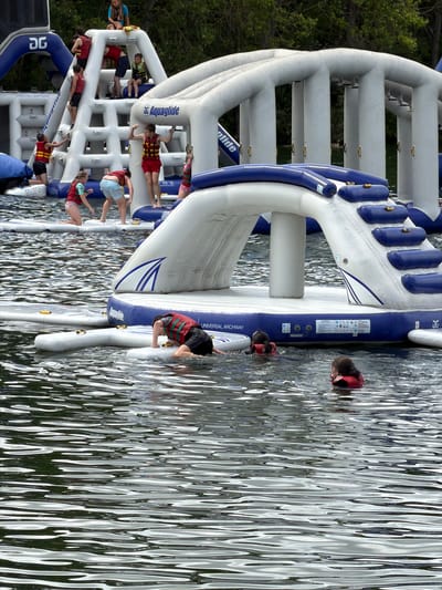 The image depicts an outdoor water park featuring large inflatable structures. The structures are white and blue, with various climbing elements, slides, and platforms. Several individuals are present, wearing life vests in various colors, including red and yellow. Some are actively engaged with the inflatable obstacles, climbing or navigating them, while others are in the water. The water is rippled, reflecting the surrounding environment. In the background, trees are visible, suggesting a natural setting. The overall scene is one of recreation and water-based activities. There is no specific indication of the city of South Ockendon, United Kingdom, within the image itself.