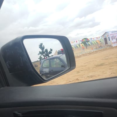 The image captures a view through a car's side mirror and the car's interior, with a scene in Wuro Ngangandi, Nigeria, visible in the mirror and beyond. In the side mirror, a dark-colored hatchback car is reflected, with text written on its rear side. Beyond the car in the reflection, a large tree with sparse foliage stands, and several people are visible standing and on a motorcycle. The ground appears to be dirt.

In the actual scene, beyond the car's interior which is in the foreground, a construction or event site is set up. Colorful triangular flags are strung across a fence. Behind the fence, a small building with a white facade and dark windows is visible, along with a banner displaying portraits of several individuals and text. There are also several tall, curved banners in shades of orange, yellow, and green. People are seen working or present at the site, including one person in an orange vest. The environment is an open, dusty area under a cloudy sky.