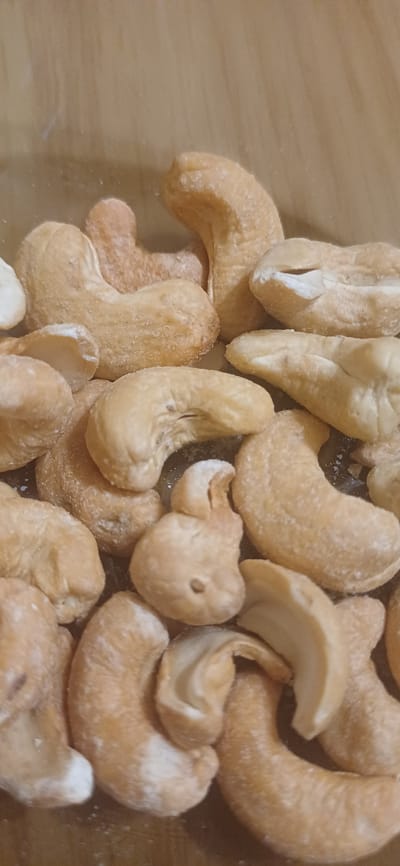 A close-up, eye-level, vertical shot captures a generous pile of roasted cashews. The cashews are a warm, golden-brown color, dusted with a light coating of salt or sugar that gives them a slightly textured appearance. They are irregularly shaped, with the characteristic kidney-like curve of a cashew nut. Some nuts are whole, while others appear to be broken or have slight imperfections. The background is a blurred, warm-toned surface, possibly wood or a similar material, providing a neutral backdrop that highlights the nuts. The lighting is soft and even, illuminating the details of the cashews without harsh shadows. The scene suggests a food-related context, likely a snack or ingredient. There are no people or animals present. The location in Zuevka, Russia is not discernible from the image.