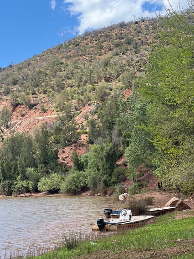 The scene, located in Ait Ouaarda, Morocco, depicts a body of brownish-green water meeting a vegetated and earthen bank. In the right foreground, two motorboats are partially beached: a larger, light-colored vessel equipped with a steering console and a black outboard motor, and a smaller, darker boat with a second black outboard motor positioned behind it. A third, smaller, light-colored flat-bottomed vessel is pulled further onto the immediate shore. A white, flamingo-shaped decorative object stands near the larger boat. The bank is covered with green grass and reeds in the immediate foreground, transitioning to denser green trees and shrubs further up the slope. A steep hillside, characterized by exposed reddish-brown soil and rocks, rises prominently in the background. Sparse to moderate vegetation, primarily small trees and scrub with green foliage, covers the hillside. A narrow, light-colored dirt track is visible traversing the mid-slope of the hill. Overhead, multiple metal electricity pylons with associated power lines are positioned along the hilltop, silhouetted against a clear blue sky featuring scattered white cumulus clouds. The scene is illuminated by natural daylight. No individuals are clearly discernible engaging in specific actions or interactions.