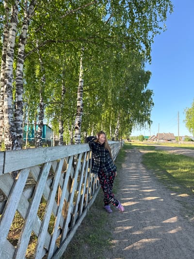 A young female with long, light brown hair stands beside a weathered wooden fence, leaning against it with her right arm bent. She wears a dark plaid shirt and dark trousers patterned with red chili peppers and white text, along with mismatched purple slippers. The fence, constructed with a latticework of gray-blue diagonal wooden slats, borders a dirt path that stretches into the distance. A prominent row of birch trees with white bark and vibrant green foliage lines the path and fence on the left side of the scene. In the background, across the path, several wooden houses with pitched roofs are visible, characteristic of rural architecture in Ust'-Silayka, Russia. Power lines on wooden poles are also present along the right side of the path. The scene is bathed in clear, warm sunlight, casting long, distinct shadows from the trees and fence across the path, indicating late afternoon or early evening. The sky is a cloudless, bright blue.