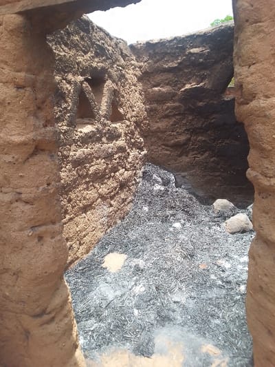 The image shows the interior of a small mud-brick building in Kwal, Nigeria, located at latitude 9.804 and longitude 8.647. The room is empty except for a layer of charred debris on the floor. There are two small openings on the wall, likely windows, and the walls are made of mud brick. The room appears to have been recently damaged by fire, as evidenced by the charred debris and the blackened walls. The sky is visible through a gap in the roof.