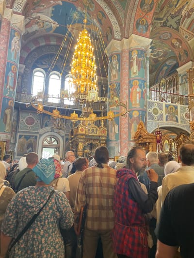 The image depicts the interior of the Holy Trinity Cathedral in Diveyevo, Russia, during a gathering. A large, ornate golden chandelier hangs prominently from the ceiling, surrounded by elaborate religious murals and architectural features. A crowd of people, mostly women, are gathered inside the cathedral, facing towards the front. Many are wearing headscarves, typical for attending religious services in Orthodox churches. The atmosphere appears to be one of reverence and participation in a spiritual event.