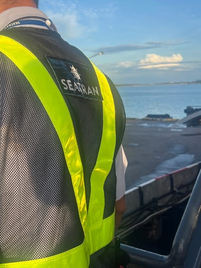 A close-up, rear-view shot focuses on the upper back and shoulder of an individual wearing a high-visibility vest. The vest is black mesh with prominent neon yellow reflective strips. A rectangular patch on the back of the vest displays a circular logo with a ship's wheel and the text "SEATRAN" underneath. A blue lanyard with the same "SEATRAN" text is visible around the person's neck, attached to a white button. The background is blurred, showing a body of water, a cloudy sky, and a dock or pier area, suggesting an outdoor setting near a waterfront. The overall scene conveys a sense of maritime activity.