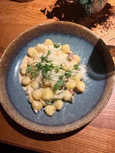 A top-down shot showcases a blue-rimmed plate filled with creamy gnocchi. The gnocchi, topped with grated cheese and garnished with fresh sprouts, are bathed in a light-colored sauce. The dish rests on a wooden table, with hints of other elements like a plant and glassware subtly visible in the background. The lighting creates shadows, adding depth to the composition.