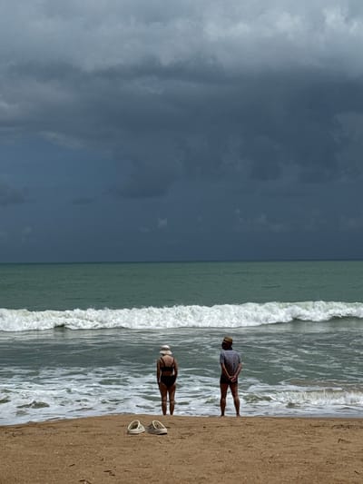 Two individuals stand on a sandy beach facing a choppy sea under a dark, overcast sky. A pair of white sandals rests on the sand in the foreground. The person on the left, viewed from behind, wears a dark bikini and a wide-brimmed hat. The person on the right, also viewed from behind, is dressed in a patterned short-sleeved shirt, dark shorts, and a straw hat. The water exhibits white, frothy waves breaking near the shore. The background is dominated by the sea and a heavy cloud cover, suggesting inclement weather. The location is the city of Ban Khuk Khak, Thailand.