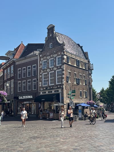 A street scene in Zwolle, Netherlands, captured on a bright, sunny day, likely in the afternoon. The image is oriented vertically, with the perspective looking upwards towards buildings and the sky. In the foreground, a woman in a light-colored outfit stands on a brick-paved street, facing the camera with a slight smile. Behind her, a storefront displaying the "CALZEDONIA" logo is visible, with mannequins and products visible through the windows. Further down the street, other people are seen, some walking and others seated outside cafes, with bicycles parked nearby. The architecture of the buildings is old and distinctive, with brick facades, white-framed windows, and tiled roofs. Some of the buildings feature ornate detailing, suggesting historical significance. The clear blue sky suggests good weather. The overall atmosphere appears relaxed and leisurely, typical of a pleasant day in a European city.