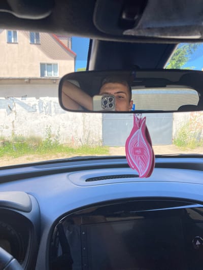 The media file shows a selfie taken in a car's rearview mirror. The subject is a person whose face is partially visible in the reflection, holding a smartphone. A pink, leaf-shaped air freshener hangs from the mirror. The car's interior, including the dashboard and part of the steering wheel, is visible. Outside the car, through the windshield and the mirror's reflection, a building with multiple windows and a red-tiled roof can be seen. The scene appears to be set on a sunny day, indicated by the bright light and clear blue sky reflected in the mirror and visible through the windshield. The general setting is urban or suburban, likely in Ustka, Poland, given the context. There is no discernible emotion conveyed by the subject. Text is visible on the air freshener, which appears to read "AROMATIC".