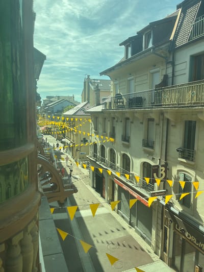 This is a medium-shot, eye-level, outdoor photograph taken on a sunny day in Évian-les-Bains, France. The image shows a street lined with buildings and decorated with yellow triangular flags strung across it.
In the foreground, on the left, a portion of a decorative architectural element with intricate carvings and green glass is visible. The street itself is paved and shows distinct shadows cast by the buildings and the flags. Several people are walking along the street, some singly and some in small groups. They are dressed in casual summer clothing.
The buildings on either side of the street are multi-story structures with awnings, balconies, and shuttered windows. Some of the buildings house shops and cafes, as indicated by signage such as "Confiserie de la Source," "Le Brummels," and "Cafe 3." The sky above is partly cloudy with streaks of white clouds. The overall atmosphere suggests a lively, festive day in a European town.