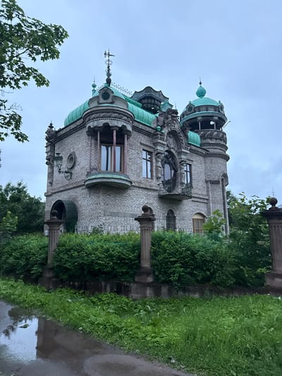 This image shows an exterior view of the Terentyev House, located in the city of Valday, Russia. The building is a unique architectural structure with a grey brick facade and multiple green domes and turrets. It features ornate details, including statues, decorative window surrounds, and a wrought-iron balcony. The house is surrounded by dense green bushes and foliage. In the foreground, a wet, muddy path with puddles suggests recent rain. The sky is overcast and grey. There are no people or animals visible in the image. The scene appears to be captured on a cloudy day.
