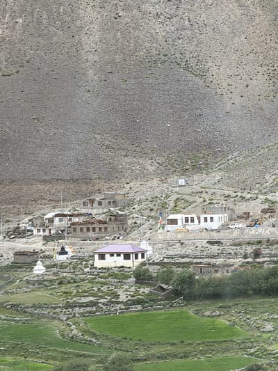The image depicts a mountainous village scene, likely in Testa, India, characterized by traditional stone and mud-brick houses clustered on a rocky slope. The architecture features flat roofs, some adorned with bundles of straw, and small windows. In the foreground, terraced green fields suggest agricultural activity, with evidence of irrigation channels. Several white stupa-like structures, common in Buddhist regions, are visible among the buildings. The background is dominated by a vast, barren, rocky mountain face, indicative of a high-altitude, arid environment. Signs of modern construction are present, including what appears to be a yellow bulldozer and white vehicles near some of the buildings. Prayer flags, displaying vibrant colors, are strung between structures, adding a touch of cultural significance to the landscape. The overall impression is one of a remote, rustic settlement nestled within a dramatic natural setting.