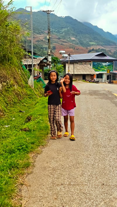 The image displays two young Asian girls on a gray asphalt road in Pung Luong, Vietnam, with a village and mountainous terrain in the background under an overcast sky.

The girl on the left wears a black short-sleeved t-shirt, brown and beige plaid pants, and light-colored sandals. Her dark hair is styled with a headband. She holds a red smartphone in her right hand and looks directly at the camera with a subtle smile.

The girl on the right wears a red short-sleeved t-shirt with light-colored text, light purple shorts, and bright yellow slide sandals. Her dark hair is shoulder-length. She raises her right hand with two fingers extended, appearing to wave or make a gesture, and smiles broadly towards the camera.

Both girls are positioned in the foreground, walking from the right towards the left side of the frame. To their immediate left, a steep embankment covered in dense green grass and some exposed soil rises. Further up the embankment, darker green foliage, including bamboo-like plants, is visible.

In the mid-ground, a village settlement extends behind the girls. It consists of multiple buildings with gray roofs, some appearing corrugated. Several utility poles with numerous power lines and equipment are present. Two cylindrical solar water heaters are mounted on a rooftop in the mid-ground. Further back on the right, a building with a dark roof features a large mural of green mountains on its facade and a sign displaying Vietnamese text. Multiple motorcycles are partially visible, parked near the village structures.

The background is characterized by large, green mountains, some sections revealing reddish-brown earth, possibly from terraced cultivation. The mountains extend into the distance under an overcast sky, which is predominantly white with hints of blue.