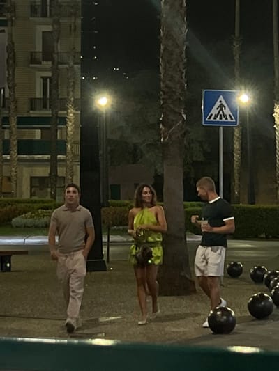 The scene depicts a nighttime outdoor urban environment in Sestri Levante, Italy, illuminated by artificial streetlights. The ground is paved, and tall palm trees are prominent throughout the area. In the background to the left, a multi-story building with balconies is visible. Green shrubbery borders the background. To the right, a blue square sign with a white pedestrian crossing symbol is mounted on a pole. Several dark, spherical objects, possibly decorative bollards or landscaping elements, are arranged on the ground. A dark, low bench or barrier is present on the far left.
Three individuals are present. On the left, a male is walking forward, wearing a light-colored polo shirt and light-colored long pants. In the center, a female is walking forward, wearing a lime-green halter-neck mini-dress and light-colored shoes; she carries a small, dark handbag and is looking towards the individual on the right. On the right, a male is standing, facing the female, wearing a dark short-sleeved shirt with white trim and light-colored shorts; he holds a clear cup with a drink and ice in his right hand. The female and the standing male appear to be engaged in a visual interaction.
