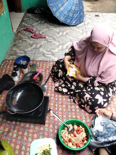 This image depicts a woman preparing food in an informal outdoor or semi-outdoor setting in Leuwiliang, Indonesia, during daytime. The primary subject, a woman wearing a pink hijab and a dark floral-patterned dress, is seated on a colorful mat. She is holding a small yellow packet that clearly shows the text "SINTA" and appears focused on its contents.
Around her, a simple cooking station is set up. A black pan rests on a portable single-burner gas stove. To the left, there's a mortar and pestle containing ground ingredients, flanked by several spice shakers and a clear mug. A blue bowl with small packets is also visible. In front of the woman, a green bowl holds a mix of ingredients, including noodles, sliced red sausages, and other items, with a metal ladle inside. A white plate with chopped green chilies and other minced ingredients is also present.
The background features a tiled floor with an intricate pattern, leading to a green-painted wall. A blue and white checkered umbrella leans against the wall, and a pair of red flip-flops are casually placed on the tiles. The bright natural light indicates it is daytime. The scene conveys a sense of domestic activity, with the woman diligently engaged in preparing a meal. A portion of another person's hand is visible in the lower right corner, suggesting shared activity.
