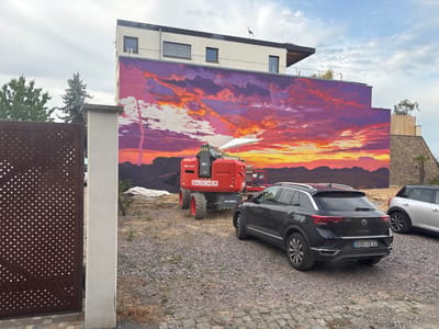 A dark grey Volkswagen T-Roc is parked on a gravel surface in front of a building with a large mural. The mural depicts a dramatic sunset with vibrant orange, purple, and pink clouds and a silhouetted tree in the foreground. The building itself is modern, with white and grey sections and several windows. A red scissor lift, branded with "BAUSCHER" and "Miete mich!", is positioned in front of the mural, suggesting ongoing work or construction. To the left of the image, a section of a dark brown, lattice-patterned gate is visible, and to the right, a silver car is partially visible. The ground is covered in gravel and some scattered debris, and the sky is partly cloudy. The scene appears to be in an urban or suburban setting, likely a construction site or the exterior of a newly developed property in Bad Dürkheim, Germany.