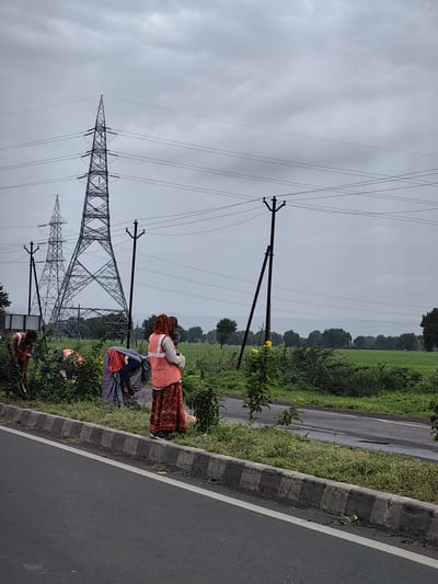 This image depicts an outdoor scene near the city of Dharangaon, India, under an overcast sky. In the foreground, a paved road with a white line is visible, bordered by a concrete curb and a grassy verge. Several individuals, likely laborers, are working on the grassy verge, tending to vegetation. They are wearing orange safety vests over traditional Indian attire. One individual in the center is wearing a patterned skirt and an orange vest, holding what appears to be a child. To the left, two other individuals in orange vests are bent over, engaged in manual labor amongst the plants. In the mid-ground, a series of tall electrical pylons and smaller utility poles with wires stretch across the landscape. The background consists of a lush green field bordered by a line of trees. The overall atmosphere is cloudy and potentially humid, suggestive of monsoon season or impending rain.