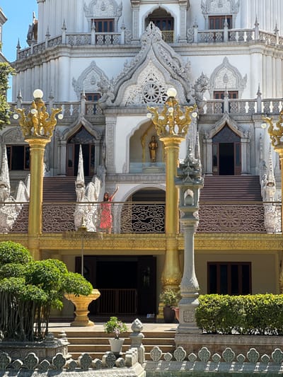 The image shows a temple, likely in Dĩ An, Vietnam, based on the architectural style. The building is predominantly white with ornate, dragon-themed decorative elements, along with gilded poles. A woman in a red dress stands on an elevated balcony, her arm raised. A patterned metal screen is visible behind her. Potted plants and trees flank the temple's entrance. The image is taken in daylight.