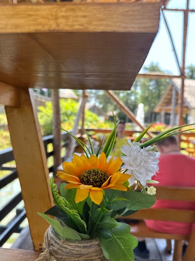 This image captures a close-up view of a decorative bouquet featuring a yellow sunflower and white flowers, placed in a glass jar wrapped with twine. The bouquet is positioned on a wooden surface, likely a table or railing, with a blurred background suggesting an outdoor setting. Beyond the flowers, wooden structures and greenery are visible, indicating a garden or outdoor seating area. In the background, two individuals are present. One person, wearing a red shirt, is seated on a wooden bench. Another person, wearing a yellow top, is standing further back. The overall scene suggests a relaxed, possibly a resort or a restaurant environment, in Indjija Municipality, Serbia. The natural light indicates it is daytime, and the clear sky hints at pleasant weather.