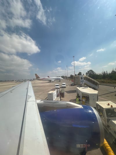 This is an eye-level, wide shot of an airport tarmac in Yehud Monosson, Israel, on a bright, partly cloudy day. The foreground is dominated by the wing and engine of an airplane, viewed from inside the cabin. The wing is a pale gray, with visible rivets and panel lines. The engine cowling is a striking metallic blue, with gray accents.

To the right of the engine, a mobile jet bridge, labeled "380 BEN GURION INTERNATIONAL AIRPORT," is connected to another aircraft. The jet bridge is gray and has visible lettering in Hebrew and English. Next to the jet bridge is a white service vehicle, and two ground crew members wearing yellow vests are visible working near a white utility box.

In the mid-ground, two Arkia airplanes are parked on the tarmac. The one closer to the viewer has a predominantly white fuselage with red and brown accents on its tail and wings. The second Arkia plane, further away, is partially obscured by the first. Several white vans and a black car are also present on the tarmac.

The background shows a line of trees and buildings in the distance, suggesting the airport is situated near a developed area. The sky is a vibrant blue, with scattered white clouds. The overall scene captures a typical airport operational environment, with aircraft, ground support equipment, and infrastructure.