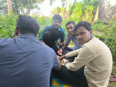 Four young men are sitting on a patterned mat in a grassy area. One man is facing away from the camera, wearing a dark blue polo shirt. Another man is wearing a teal polo shirt and is looking directly at the camera. The third man is wearing a light blue and white striped shirt and is looking slightly to the right. The fourth man is wearing a light beige shirt and is looking directly at the camera. All men are sitting on the ground and appear to be chatting. They are surrounded by lush greenery, with large palm trees in the background. The scene is located in Kalmunai, Sri Lanka, at the coordinates 7.408 latitude and 81.763 longitude.